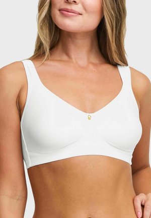 Vrouw die een witte naadloze beha draagt met brede bandjes en een klein gouden decoratief hangertje middenvoor, tegen een effen achtergrond.
