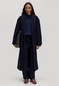 Lange marineblauwe wollen jas met een dubbelgeknoopte voorkant, gecombineerd met een gestructureerde marineblauwe trui, wijde broekspijpen en versierde bordeauxrode schoenen.