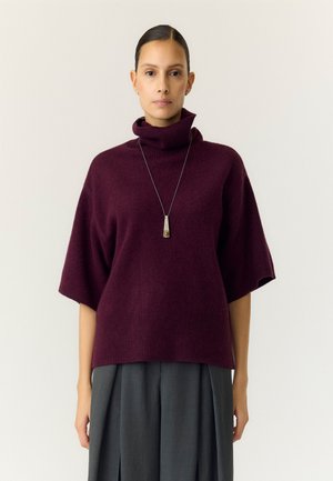 Femme aux cheveux foncés en chignon portant un pull à col roulé violet avec des manches aux coudes et un long collier pendentif.