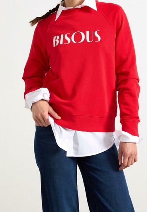 Person iført en rød sweatshirt med teksten "BISOUS", lagd over en hvid skjorte med krave og opsmøgede ærmer samt blå jeans.