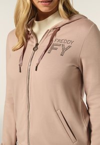 Felpa rosa chiaro con zip, tasche frontali, cappuccio con lacci e dettagli in strass che formano la scritta "FREDDY FY" sul petto. Texture liscia.