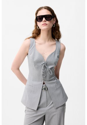 Kobieta ubrana w szarą bezrękawnikową kamizelkę z wiązaniem, dopasowane spodnie, czarny pasek, duże czarne okulary przeciwsłoneczne i kolczyki koła na białym tle.