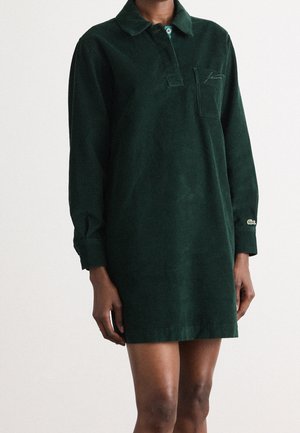 Dunkelgrünes Cordkleid mit langen Ärmeln, Kragen, aufgesetzter Fronttasche und gesticktem Logo, getragen von einer Person vor einem einfarbigen Hintergrund.