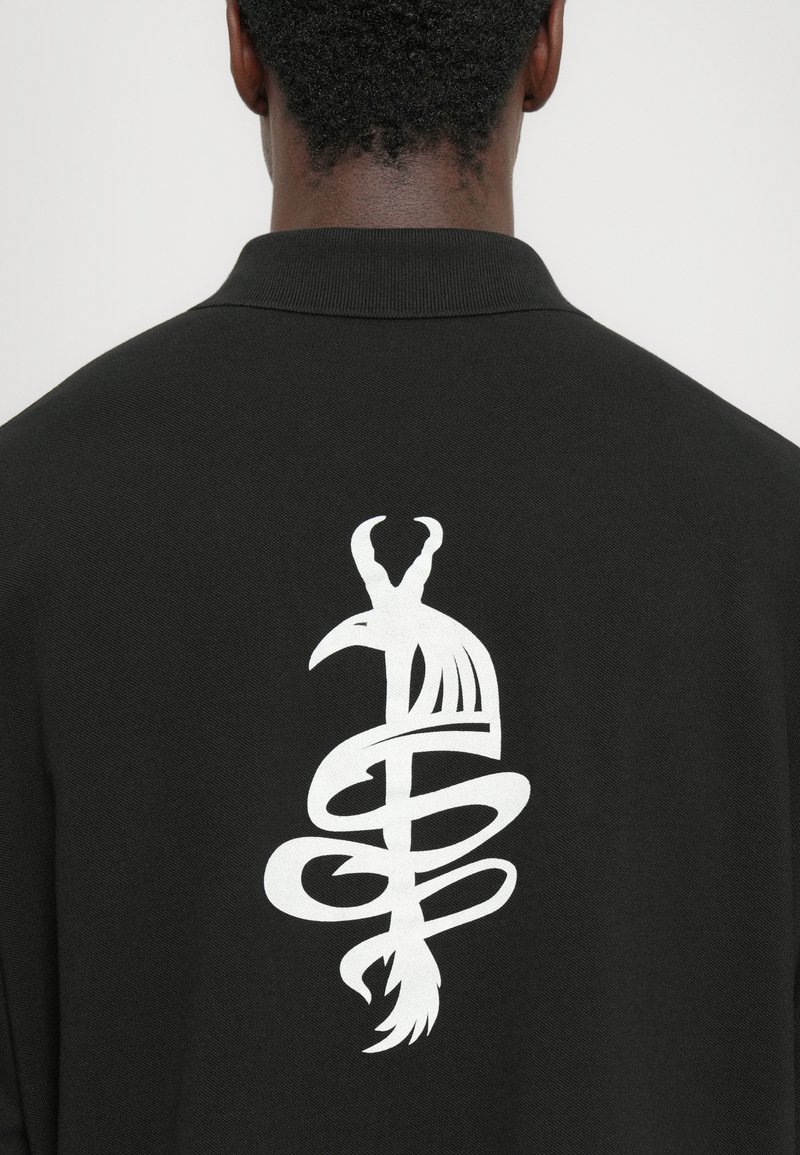 Polo nero con una grande grafica bianca di un uccello intrecciato con un serpente sul retro. Texture liscia e colletto classico.