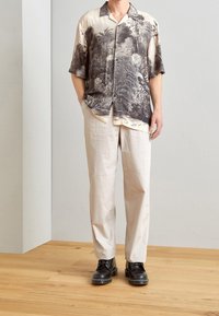 Uomo che indossa stivali neri, pantaloni beige dritti e una camicia a maniche corte con bottoni con una stampa botanica in bianco e nero.