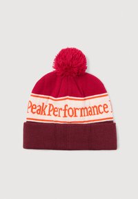 Sarkans adīts beanie ar pom-pomu, ar baltu un oranžu "Peak Performance" logo jostu ap centrā un ar piedurkni apakšā.