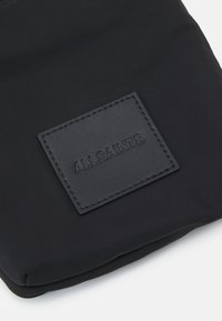 Czarna tekstylna torba o gładkiej powierzchni. Posiada prostokątną skórzaną łatkę o fakturze, wytłoczoną logo "ALLSAINTS".