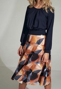 Femme portant un top noir à manches longues avec une taille haute, associé à une jupe de genoux à motifs géométriques dans des tons orange, noir et bleu.
