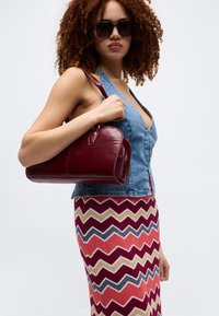 Γυναίκα με σγουρά μαλλιά που φοράει γυαλιά ηλίου, αμάνικο τζιν τοπ και πολύχρωμη φούστα με ζιγκ-ζαγκ, κρατώντας μια μπορντό δερμάτινη τσάντα.