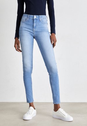 Persoon die lichtblauwe skinny jeans, witte sneakers en een zwarte top met lange mouwen draagt, staand op een plain vloer.