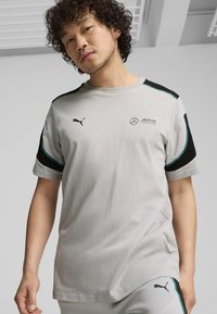 Vaaleanharmaa lyhythihainen paita mustilla yksityiskohdilla. Sisältää Puma- ja Mercedes-Benz -logot, kaarevan helman ja kontrastiväriset ompeluyksityiskohdat.