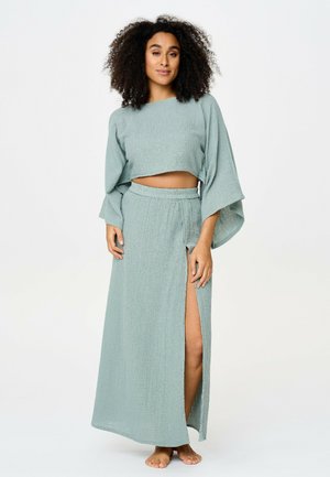 Žena s kučeravými vlasmi oblečená v svetlomodrom dvojdielnom outfite so širokými rukávmi a maxi sukňou s vysokým rozparkom, stojaca bosá na bielom pozadí.