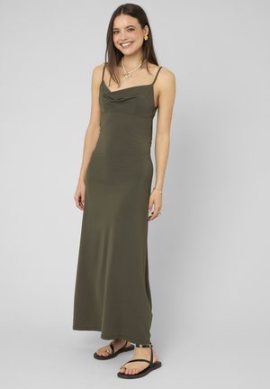 Femme souriante, vêtue d'une robe maxi sans manches vert olive à fines bretelles, sandales noires, collier étoile de mer et boucles d'oreilles en forme de fleur.