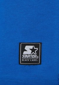 Mėlyna medžiaga pasižymi tekstūruotu mezgimo raštu. Juodas stačiakampis etiketė rodo "STARTER" ir "BLACK LABEL" baltomis raidėmis su žvaigždutės akcentu.