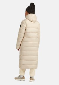 Langer beiger Daunenmantel mit horizontaler Steppung, Kapuze und einem schwarzen Logo-Patch am Ärmel. Kombiniert mit beigen Fleece-Hosen und Sneakers.