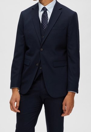 Mężczyzna ubrany w ciemnogranatowy garnitur z dopasowaną kamizelką, jasnoniebieską koszulą i granatowym krawatem w małe różowe kropki, stojący na jednolitym tle.