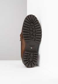 Botte en cuir marron avec une semelle en caoutchouc noir robuste, présentant des motifs profonds et un logo. Détail de fermeture éclair sur le côté pour un enfilage facile.