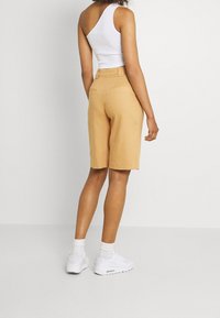 Tan linen shorts with a relaxed fit, featuring two back pockets. Paired with a white one-shoulder top and white sneakers.