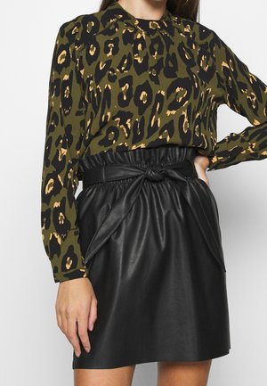 Femme portant un chemisier à imprimé léopard vert et noir, rentré dans une jupe en cuir noire taille haute avec une ceinture nouée.