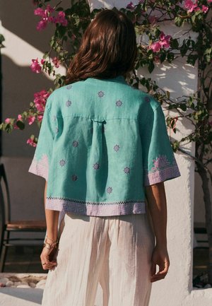 Person with brown hair wearing a turquoise short-sleeve shirt with sun patterns and white pants, standing in front of a flowering plant and white wall.