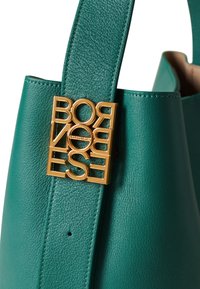 Teal leather handbag with a gold logo detail. Features a structured shape and a smooth texture. Accented with a stitched strap.