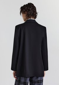 Blazer nero su misura con un design strutturato, maniche lunghe e una texture liscia. Presenta una sola apertura posteriore e dettagli minimi.