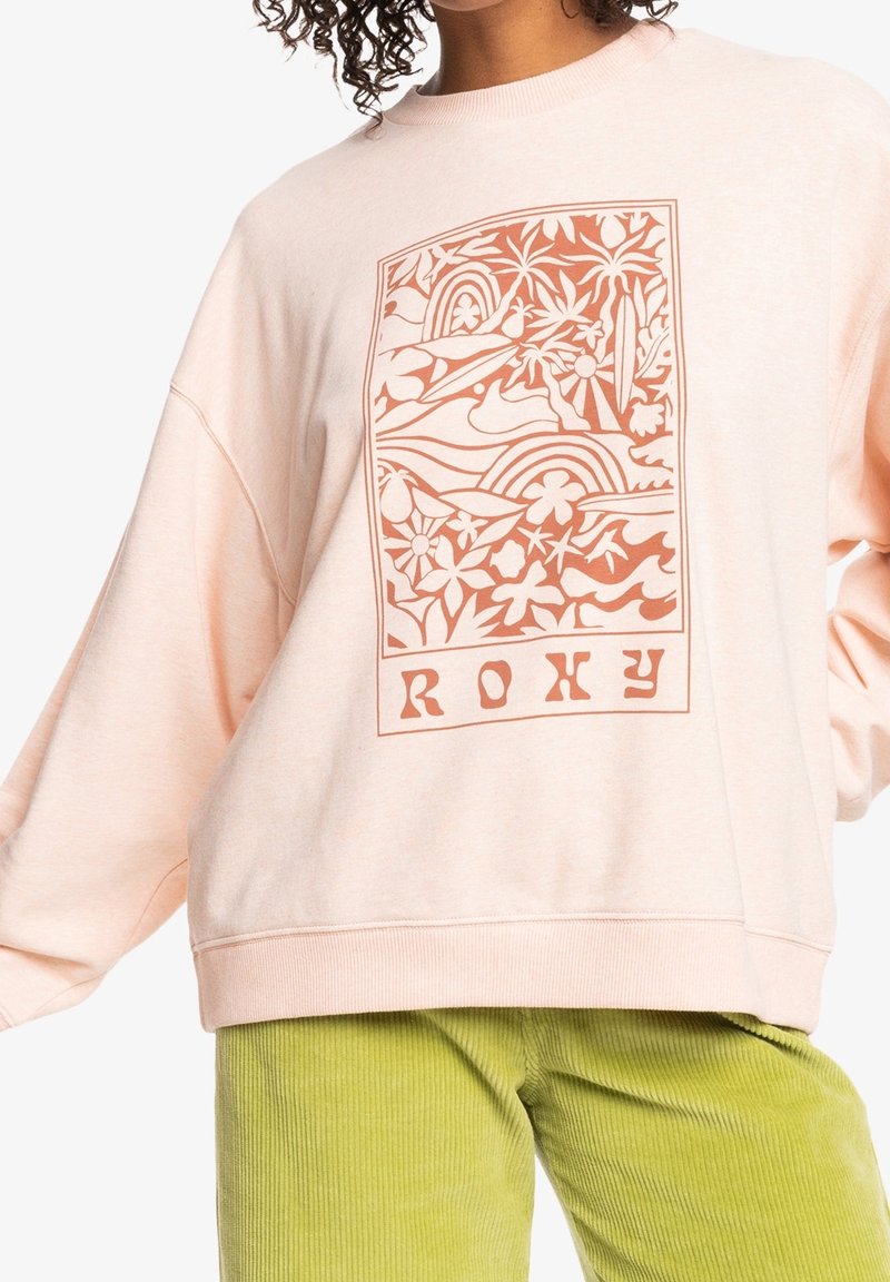 Personne portant un sweat-shirt rose pâle avec un motif floral orange et des vagues, accompagné du texte "ROXY", assorti à un pantalon en velours côtelé vert clair.