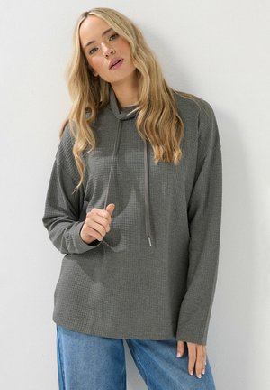 Pullover grigio in maglia waffle con cappuccio e cordoncino, caratterizzato da maniche lunghe e un taglio rilassato. Abbinato a jeans azzurri chiari.