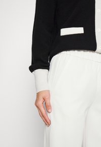 Cardigã preto com detalhes brancos, apresentando um fecho frontal com botões e punhos canelados, combinado com calças brancas de perna larga. Textura suave.