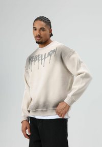 Licht beige sweatshirt met een verloop van licht naar donker aan de zoom. Heeft een "REBELLION"-grafiek in een druipende stijl, met textuurstof.