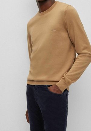 Sandfarbener Strickpullover mit rundem Halsausschnitt und gerippten Bündchen. Mit dezentem Branding auf der Brust; kombiniert mit dunklen, strukturierten Hosen.
