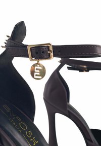 Sandal a tacco alto marrone con cinturino alla caviglia stretto, fibbia dorata e un ciondolo metallico. Presenta una texture liscia e una forma a punta.