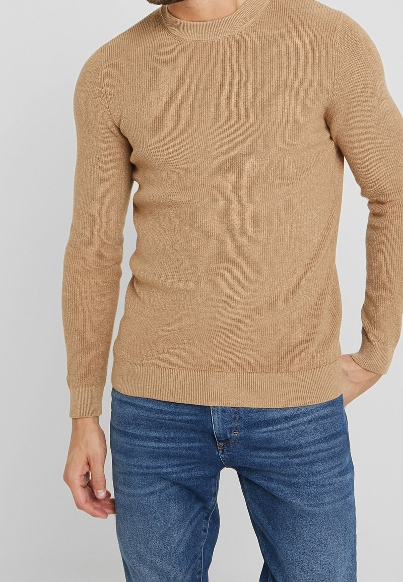 Pull beige à côtes avec de longues manches et un col rond, présentant une texture douce et une coupe ajustée, associé à un jean bleu.