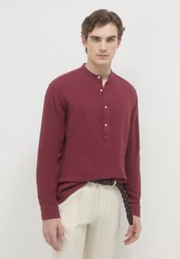 Giovane uomo che indossa una camicia strutturata a maniche lunghe bordeaux con bottoni e pantaloni beige con una cintura intrecciata marrone scuro.