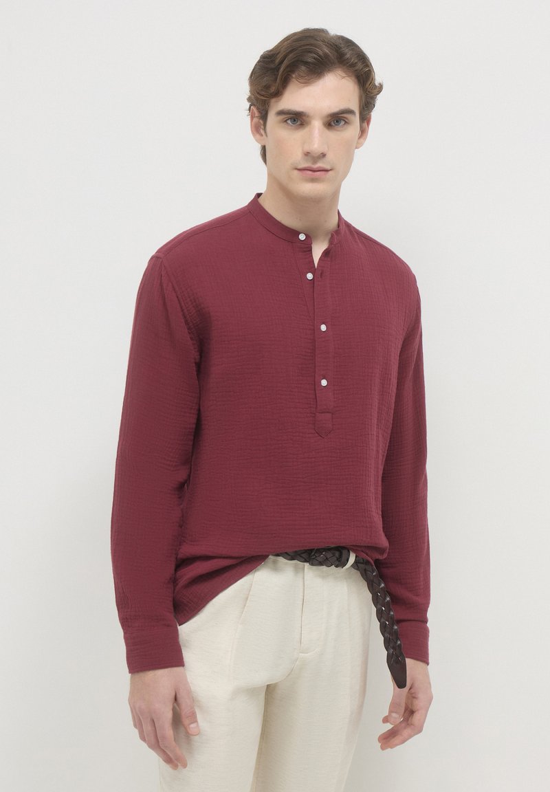 Giovane uomo che indossa una camicia strutturata a maniche lunghe bordeaux con bottoni e pantaloni beige con una cintura intrecciata marrone scuro.