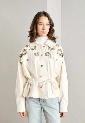 Giubbotto di jeans bianco con chiusura a bottoni dorati, design floreale ricamato sulle spalle, vita con cintura e vestibilità rilassata; abbinato a jeans blu chiaro.