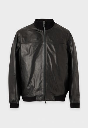 Un homme porte une veste en cuir foncée zippée, un pantalon noir et des baskets noires sur un fond clair et uni.