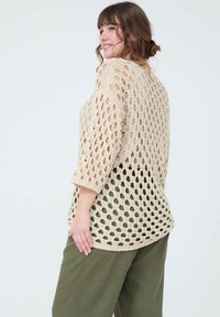 Haut en crochet beige avec un motif ajouré, coupe décontractée, manches trois quarts et ourlet arrondi, associé à un pantalon vert olive.