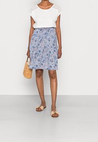 Lightweight white top with crochet detailing, paired with a blue floral skirt. Model holds a woven bag, wearing light-colored slip-on shoes.