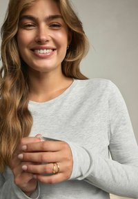 Lys grå ribbet langærmet top med rund hals. Har tætsiddende manchetter og en glat tekstur. Modellen holder hænderne sammen og viser en ring.
