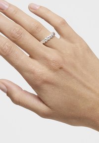 Srebrny pierścionek o nieregularnych, zaokrąglonych kształtach, noszony na dłoni o lekko teksturowanej skórze. Neutralne tło podkreśla design pierścionka.