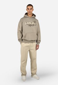 Beżowa bluza z kapturem z kwiatowym wzorem na klatce piersiowej, z kieszenią kangurką i ściągaczami na rękawach. Zestawiona z jasnobeżowymi spodniami i białymi sneakersami.