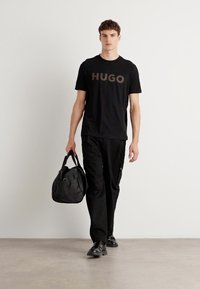 Melnas īsām piedurknēm T-krekls ar "HUGO" brūnā krāsā, apvienots ar melnām kravas biksēm un melnu somu. Modelis valkā melnus apavus. Vienkāršs fons.