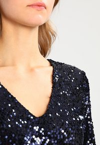 Top nero con paillettes con scollatura a V. Presenta numerose paillettes tonde e lucide, creando una superficie testurizzata e riflettente.