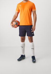 Man i orange Puma-tröja, marinblå shorts, vita Puma-strumpor och fotbollsskor som håller en orange och svart fotboll mot en enkel bakgrund.