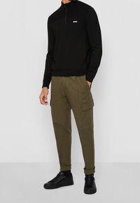 Black knit pullover with a half-zip and white logo, paired with olive green cargo pants featuring side pockets and black sneakers.
