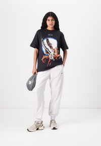 T-shirt graphique noir mettant en avant une figure ailée, associé à un pantalon de survêtement blanc et un sac Nike gris. Les chaussures sont beiges avec des accents foncés.