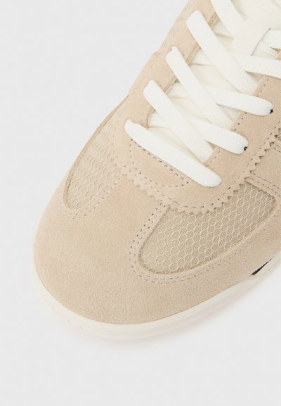 Μπεζ μύτη αθλητικού παπουτσιού με πάνελ από σουέτ και δίχτυ, λευκά κορδόνια και ανάγλυφη ραφή σε λευκό φόντο.