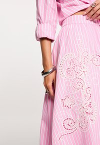 Mano de mujer descansando sobre una falda rosa con rayas blancas y bordado floral blanco, con pulsera y anillo plateados, y una camisa con las mangas remangadas.