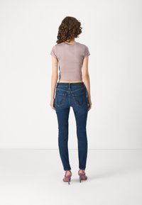Camiseta corta de color lila, combinada con unos jeans ajustados de color azul oscuro que tienen dos bolsillos traseros. El modelo lleva sandalias de tacón moradas, vistas desde la parte trasera.